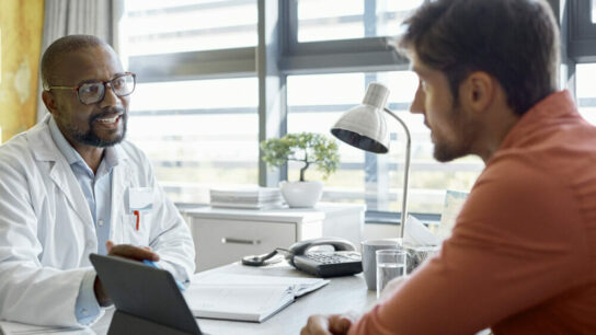 Male-doctor-and-young-male-patient-sitting-at-desk-in-a-clinic-having-a-discussion-while-using-a-digital-tablet.